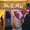 Bride and groom dancing closely indoors with illuminated 'Mr & Mrs' sign behind them.